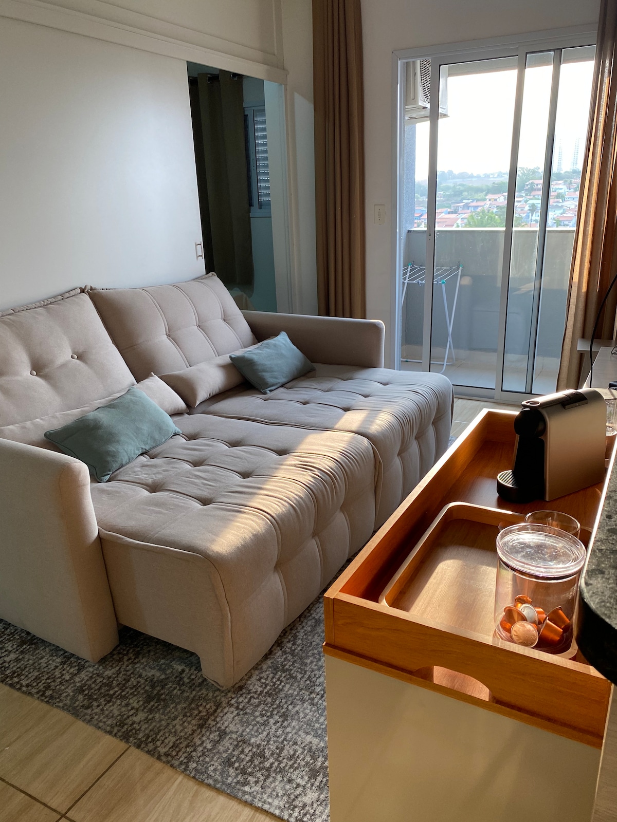 A light-colored sofa with tufted design is positioned in a cozy living area, accompanied by two soft cushions. A wooden tray on a counter holds a beverage and a snack. Sunlight streams in from a nearby glass door that opens to a balcony.