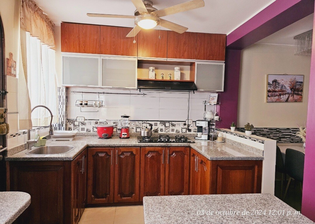 A modern kitchen is presented, featuring wooden cabinetry and a granite countertop. Essential appliances are arranged, including a gas stove, coffee maker, and various kitchen utensils. A ceiling fan enhances airflow, while a dining area is visible in the background, distinguished by a neutral color palette.