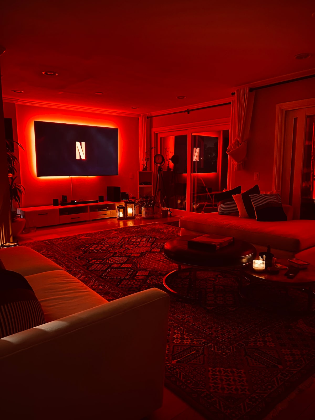 A living area is illuminated by soft red lighting, featuring a large screen mounted on the wall displaying the Netflix logo. Comfortable seating arrangements include a sectional sofa and cushions arranged invitingly. A coffee table with decorative items and candles adds warmth to the space.