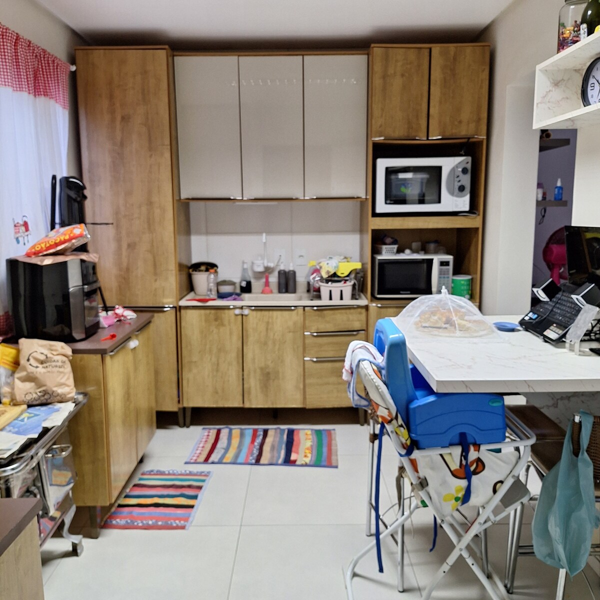 A modern kitchen is designed with wooden cabinetry and contains various appliances, including a microwave and coffee maker. A marble countertop offers preparation space, while a dining table with high chairs is positioned nearby, complemented by colorful rugs on the floor.