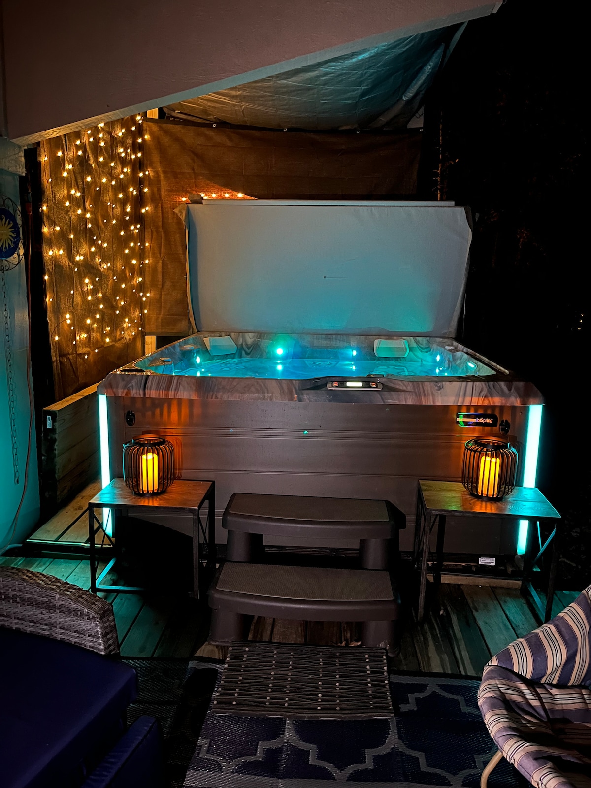 A premium saltwater hot tub is illuminated with soft blue and green lights, creating a calming ambiance. Two lanterns sit on side tables, and ambient string lights are visible in the background, enhancing the relaxing outdoor space.