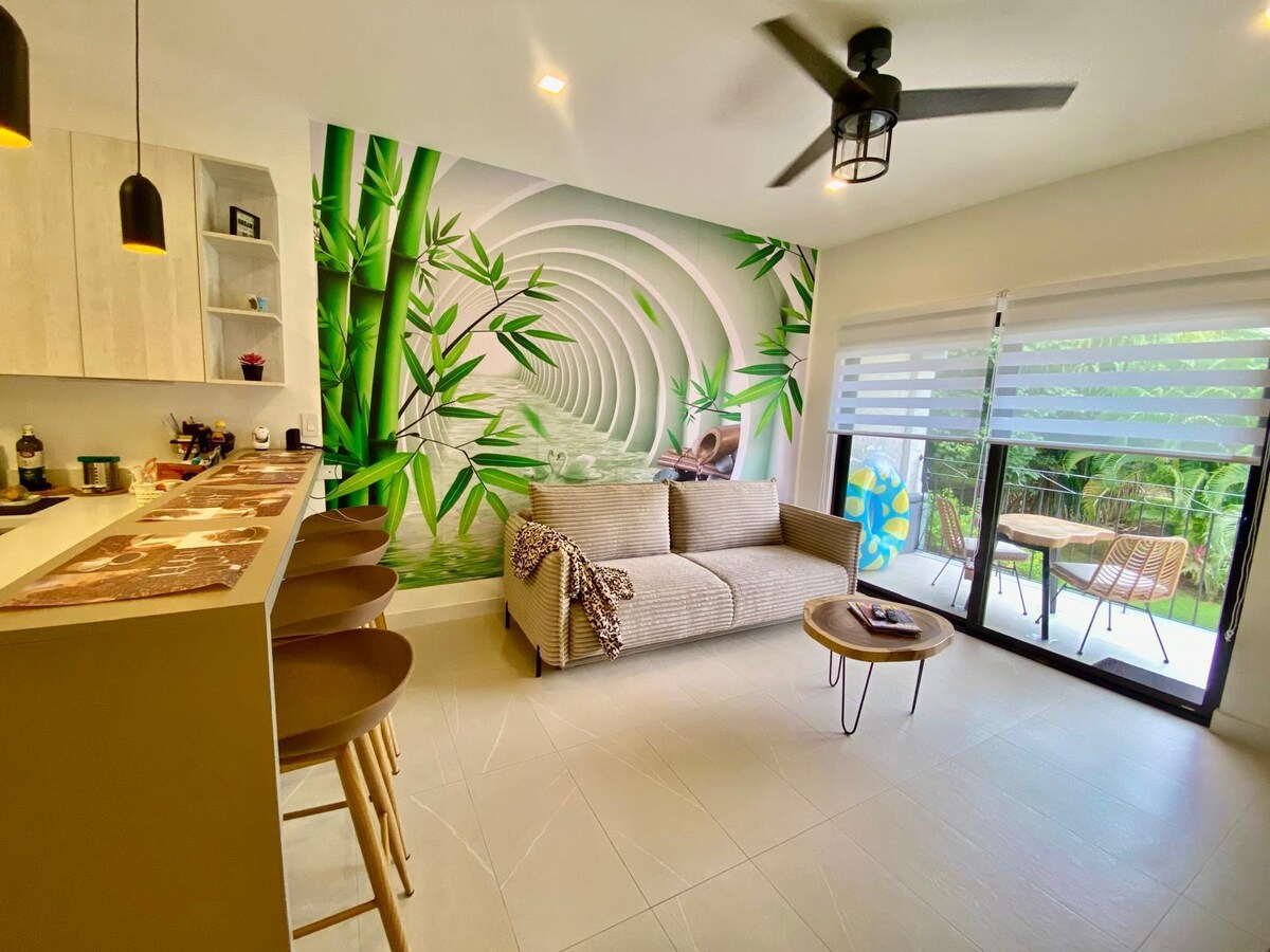 A spacious living area features a modern sofa adorned with a textured throw, complemented by a striking bamboo mural on the wall. Natural light enters through large sliding glass doors, leading to a small outdoor patio surrounded by greenery.