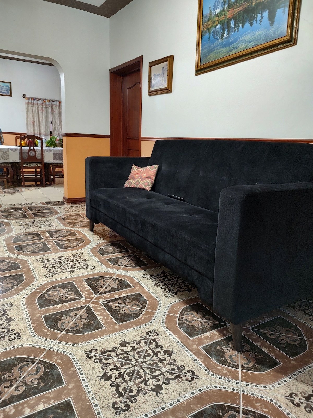 A black couch with a decorative pillow is positioned in a corner of a spacious living area. The floor is covered with patterned tiles, and a dining area with wooden furniture is visible in the background. Walls are painted in warm tones, complemented by framed artwork.