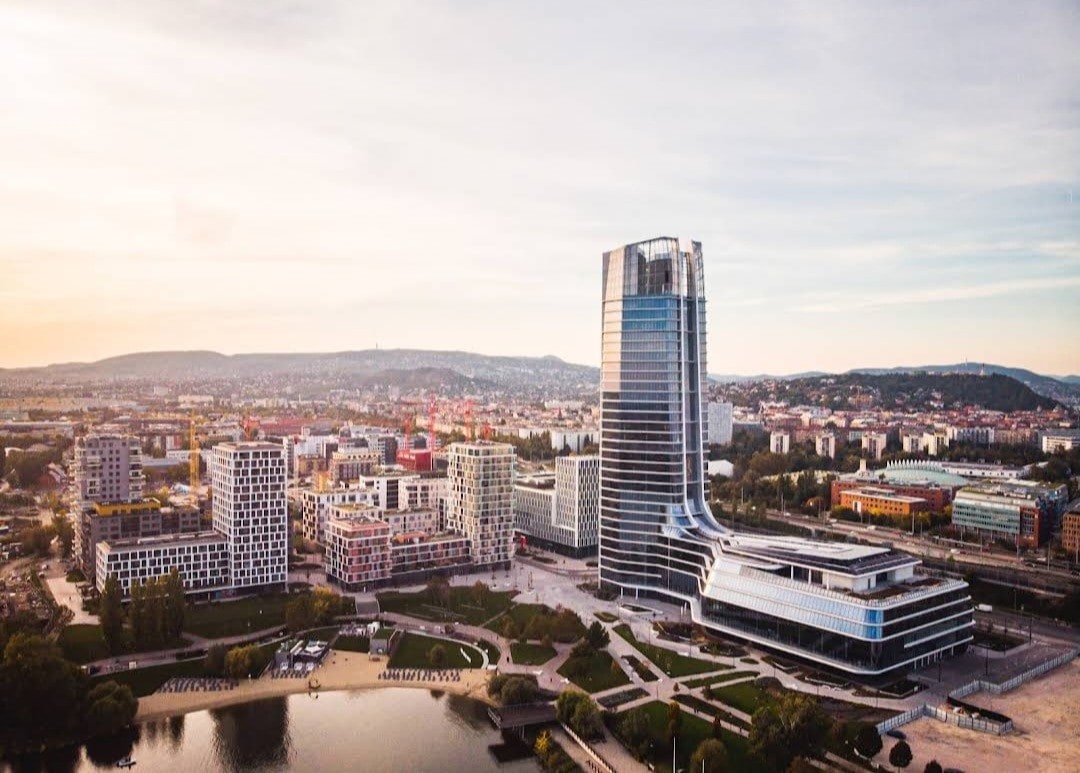 A panoramic view captures a modern skyline featuring a distinctive tall building with a sleek design. Surrounding structures exhibit varied architectural styles, while a serene body of water and green spaces are visible in the foreground, contributing to the urban landscape.
