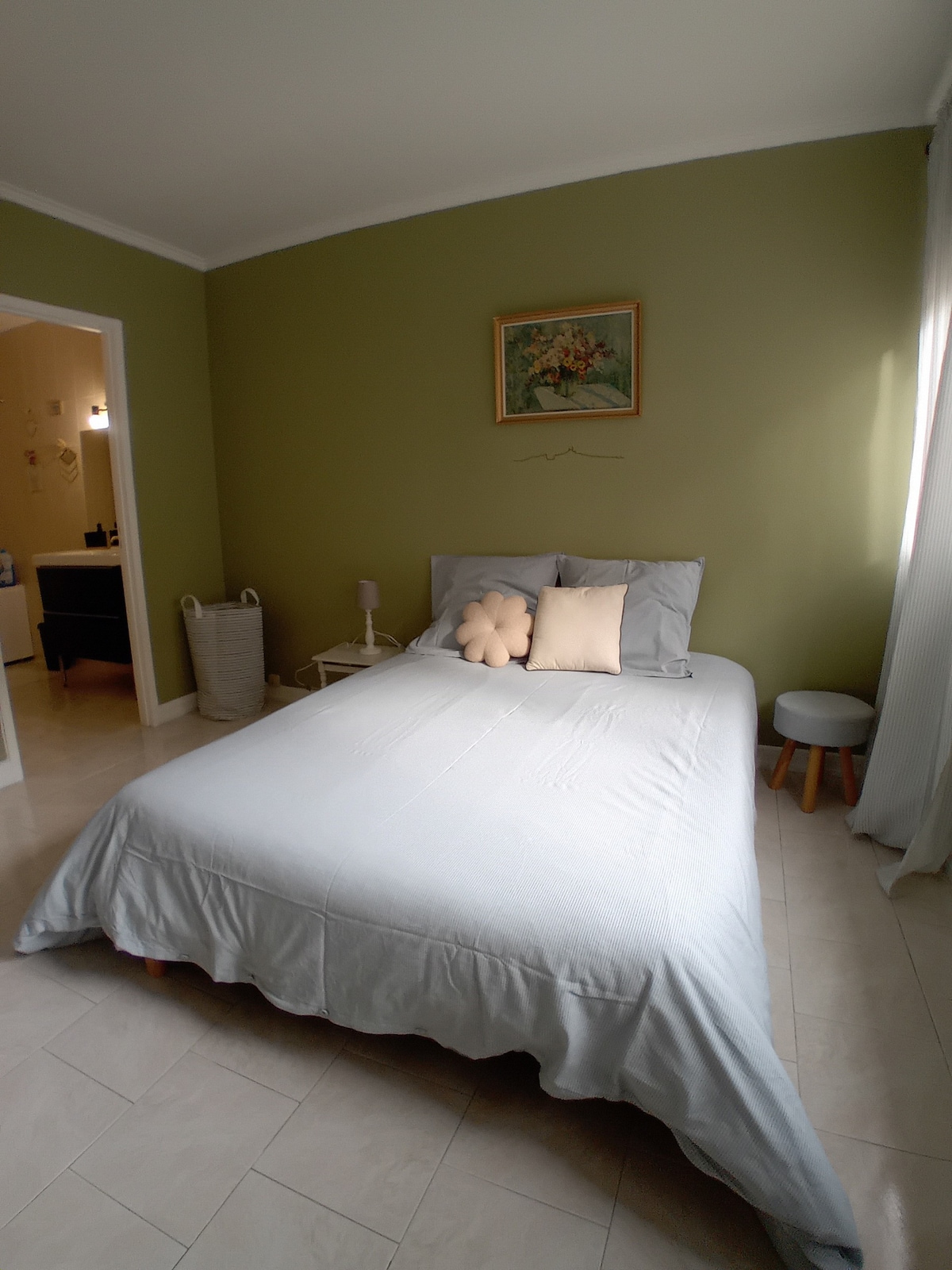 A serene sleeping area is presented with a soft gray bedspread covering a spacious bed. Accented by light green walls, a beige decorative pillow and a simple piece of artwork are positioned on the wall above. A small stool and a lamp are nearby, with natural light filtering through sheer curtains.
