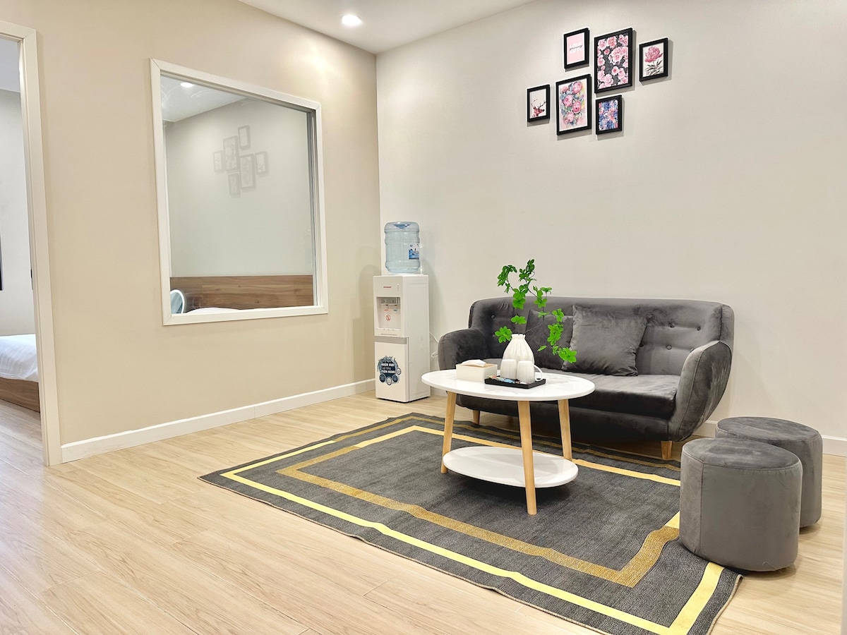 A light-filled living area is presented with a gray sofa, accompanied by a round coffee table atop a patterned area rug. Decorative wall art features floral designs, while a water dispenser and a mirror enhance the inviting atmosphere.