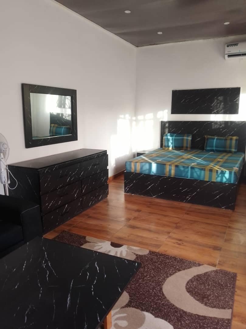 A modern bedroom showcases a bed with a blue and yellow plaid design, accompanied by matching pillows. A sleek dresser and a small table in dark marble pattern complete the space. A large mirror reflects natural light, enhancing the bright and clean atmosphere.