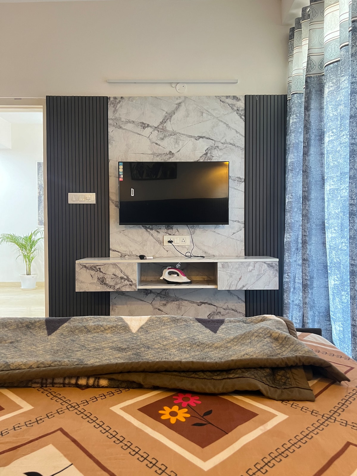 A flat-screen television is mounted on a marbled accent wall, accompanied by a sleek wall shelf. The room features a comfortable bed with a patterned blanket, and soft curtains frame the window, allowing natural light to create a warm ambiance.