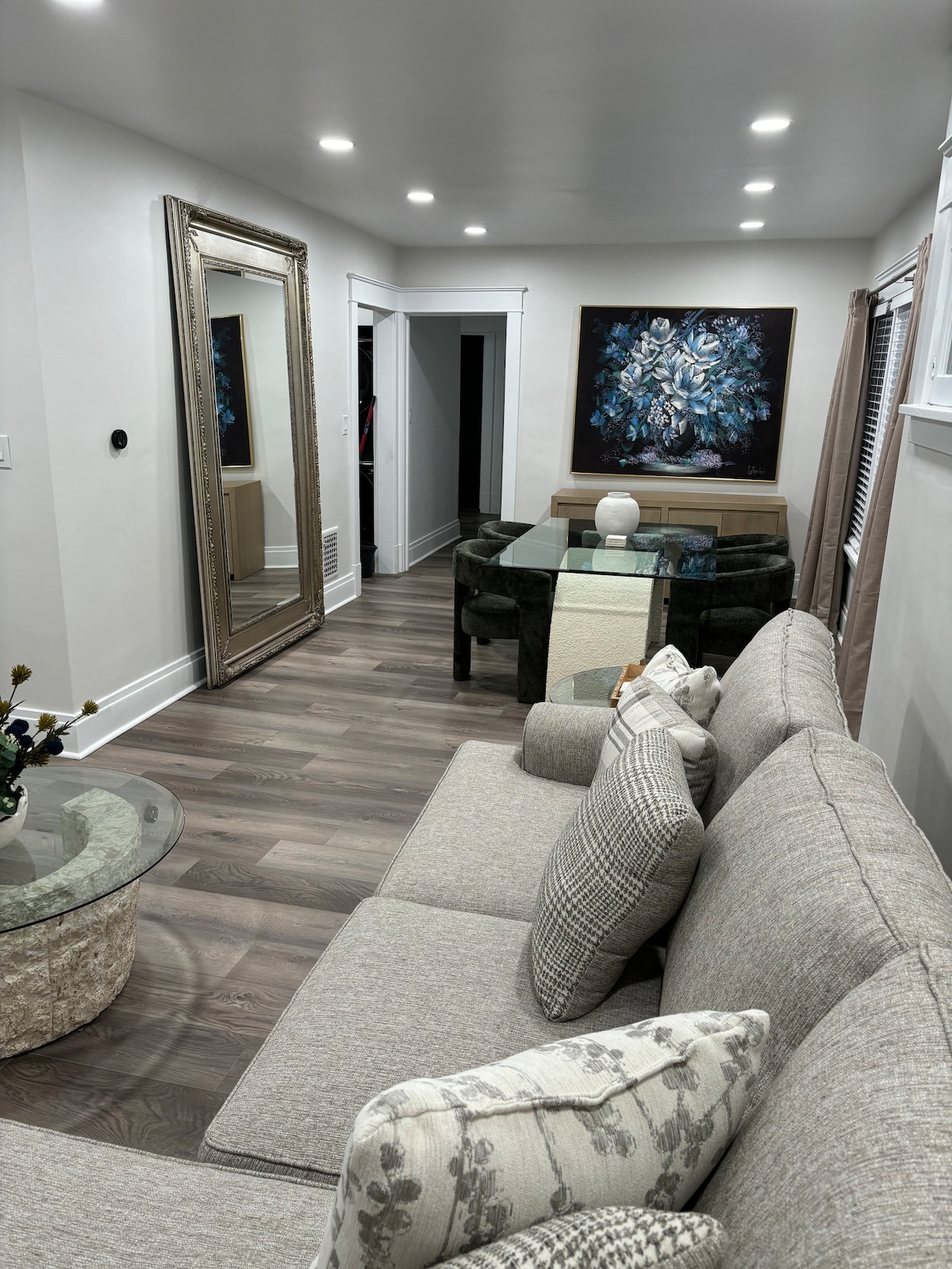 A modern living area features a beige sofa adorned with patterned pillows. A round glass coffee table sits beside it. A large decorative mirror reflects the light, while a dark dining table is visible in the background, complemented by an abstract wall art piece.