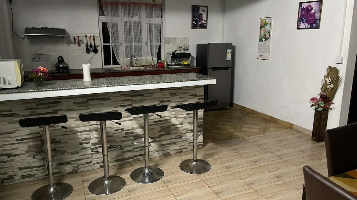 A modern kitchen area features a sleek countertop with four bar stools in front. Framed artwork adorns the walls, adding a touch of interest. Natural light filters through a window, illuminating the space that includes essential appliances and a well-organized layout.