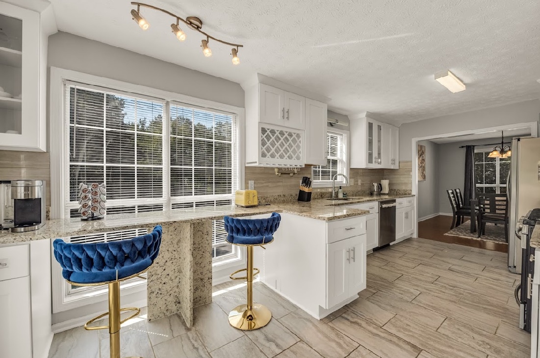 A modern kitchen features sleek white cabinetry and elegant granite countertops, with ample natural light streaming in through large windows. Two blue bar stools are positioned at the counter, providing a casual dining space. The dining area is visible in the background, enhancing functionality.