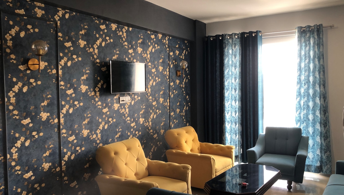 A well-lit living area features a pair of yellow armchairs facing a black coffee table. A blue accent chair is positioned nearby. The wall is decorated with a floral pattern, and large windows are dressed with sheer blue and dark curtains, allowing natural light to filter in.