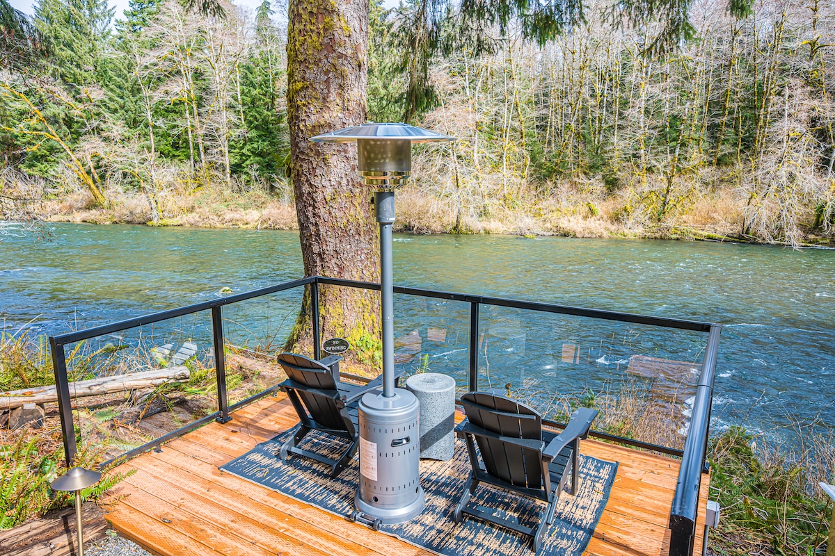 A serene outdoor seating area is presented, equipped with two reclining chairs positioned on a wooden deck overlooking the Sol Duc River. A patio heater is centrally located, providing warmth, while wild greenery and trees frame the natural setting, enhancing the peaceful ambiance.