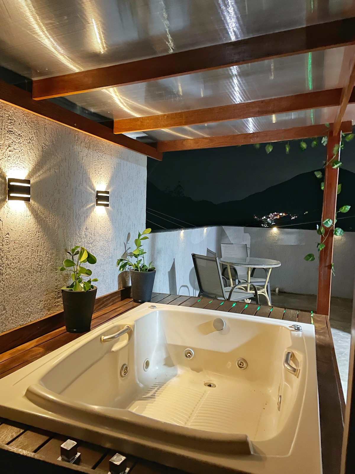 The outdoor terrace features a private jacuzzi under a wooden pergola, enhanced by soft lighting. Two potted plants are placed nearby, and a small table with chairs offers a comfortable spot for relaxation. The view beyond showcases a quiet, serene nighttime landscape.
