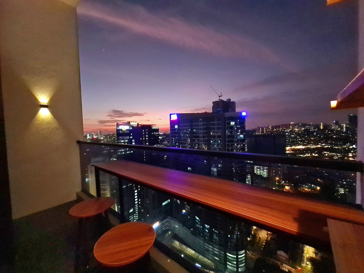 The balcony offers a sweeping view of the city at dusk, with a wooden bar table and two stools positioned for relaxation. Soft lighting highlights the space, creating an inviting ambiance as the sky transitions to evening hues.