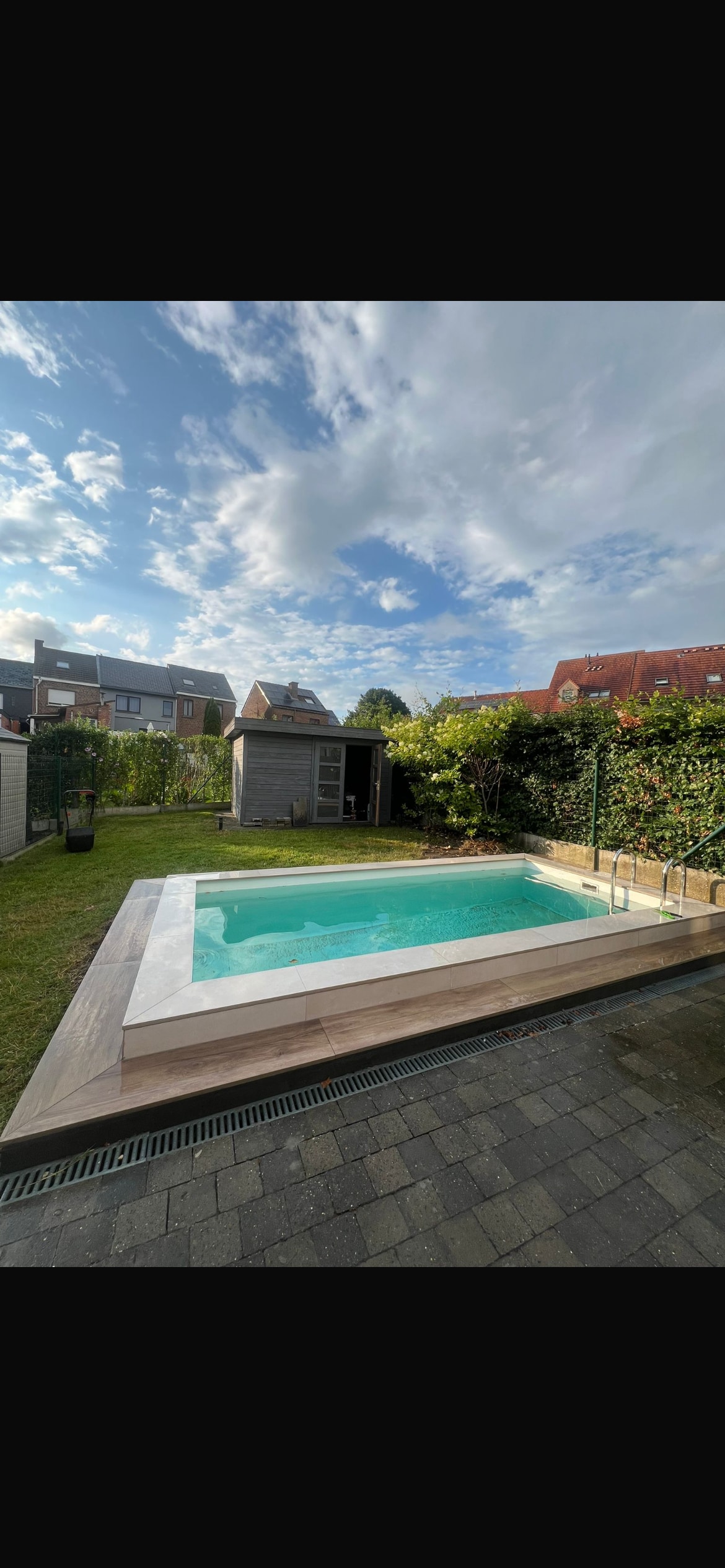 A private garden features a swimming pool surrounded by wooden decking. Lush greenery offers privacy, while a small shed is visible in the background. The sky is partly cloudy, allowing natural light to illuminate the space.