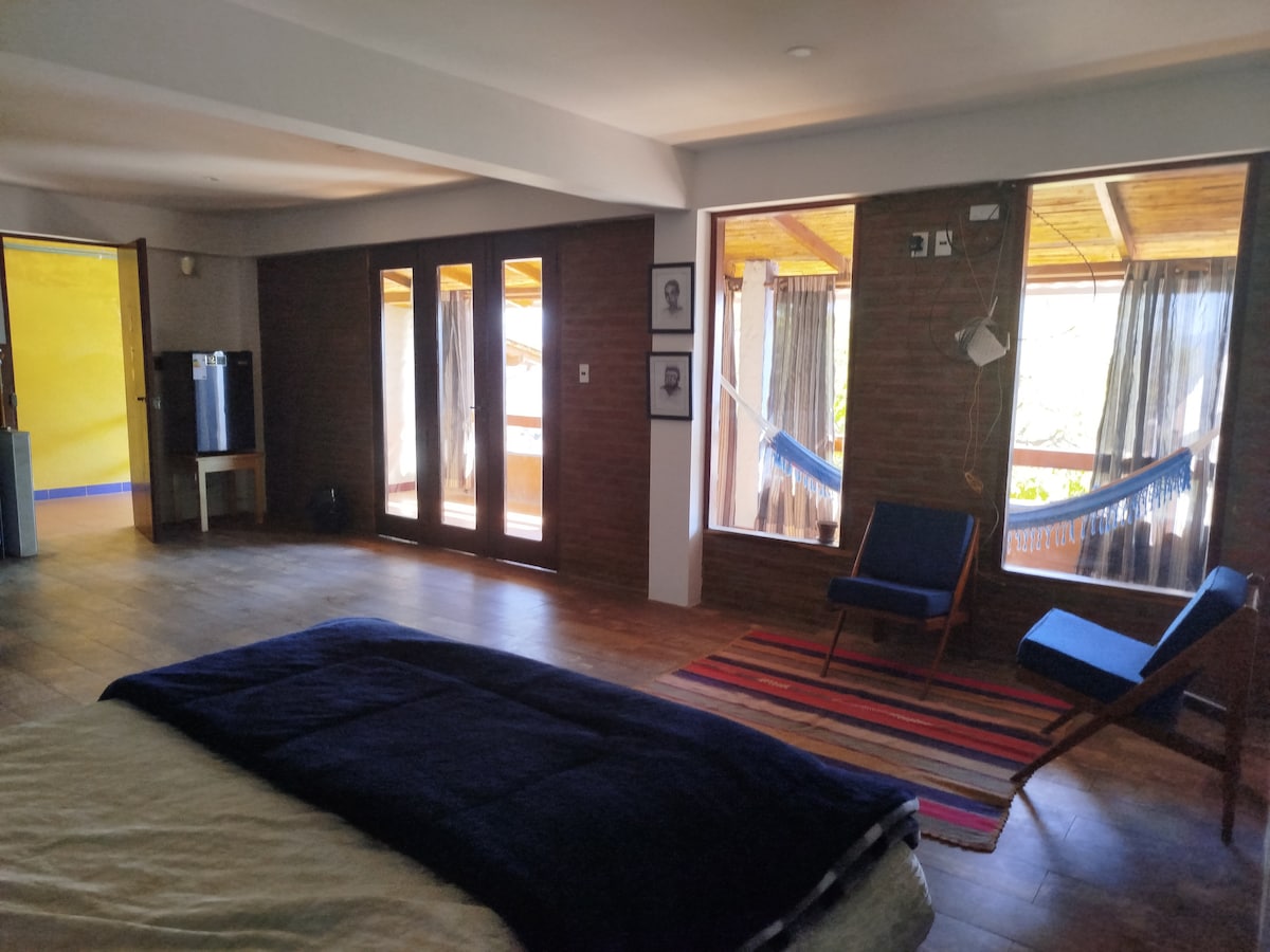 A spacious bedroom is complemented by large windows, allowing ample natural light. Comfortable chairs and a hammock create inviting relaxation spots. Exposed brick walls add texture, while a patterned rug brings warmth to the wooden floor. A small kitchenette is visible in the background.
