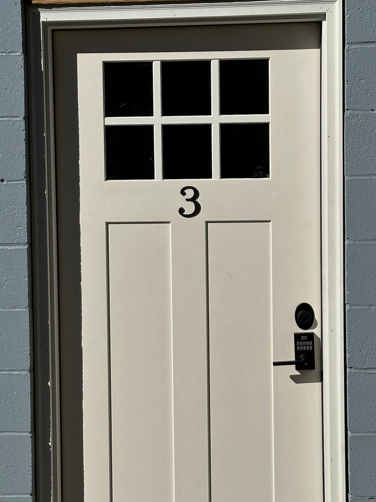 A clean, white door marked with the number three stands against a gray wall. The door features a window with four panes at the top and a modern keypad lock is installed on the right side.
