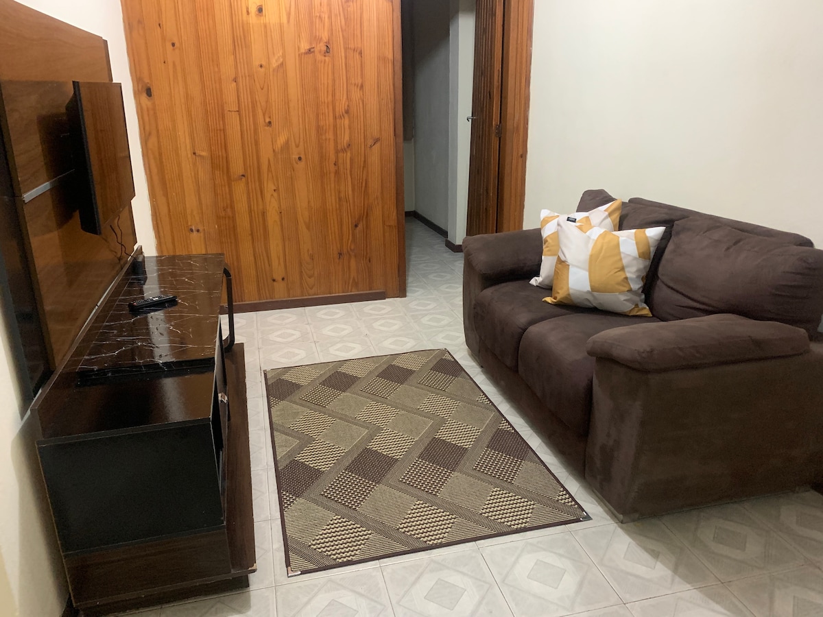 A cozy living area is presented, featuring a brown upholstered sofa adorned with decorative pillows. A flat-screen television is mounted on a wooden panel, complemented by a dark wood media console. A patterned area rug enhances the space, with light-colored tiled flooring adding brightness.