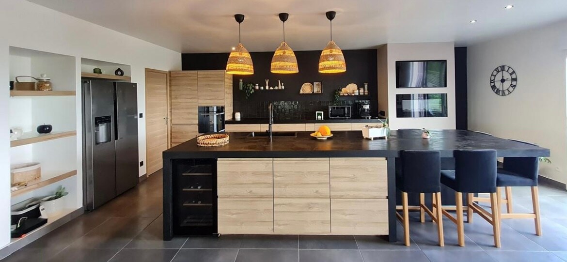 A modern kitchen features a large island with a dark countertop and wooden cabinetry. Stylish pendant lights hang above the island, while a stainless steel refrigerator and built-in appliances complement the space. Black and white accents add contrast, creating a functional and inviting cooking area.