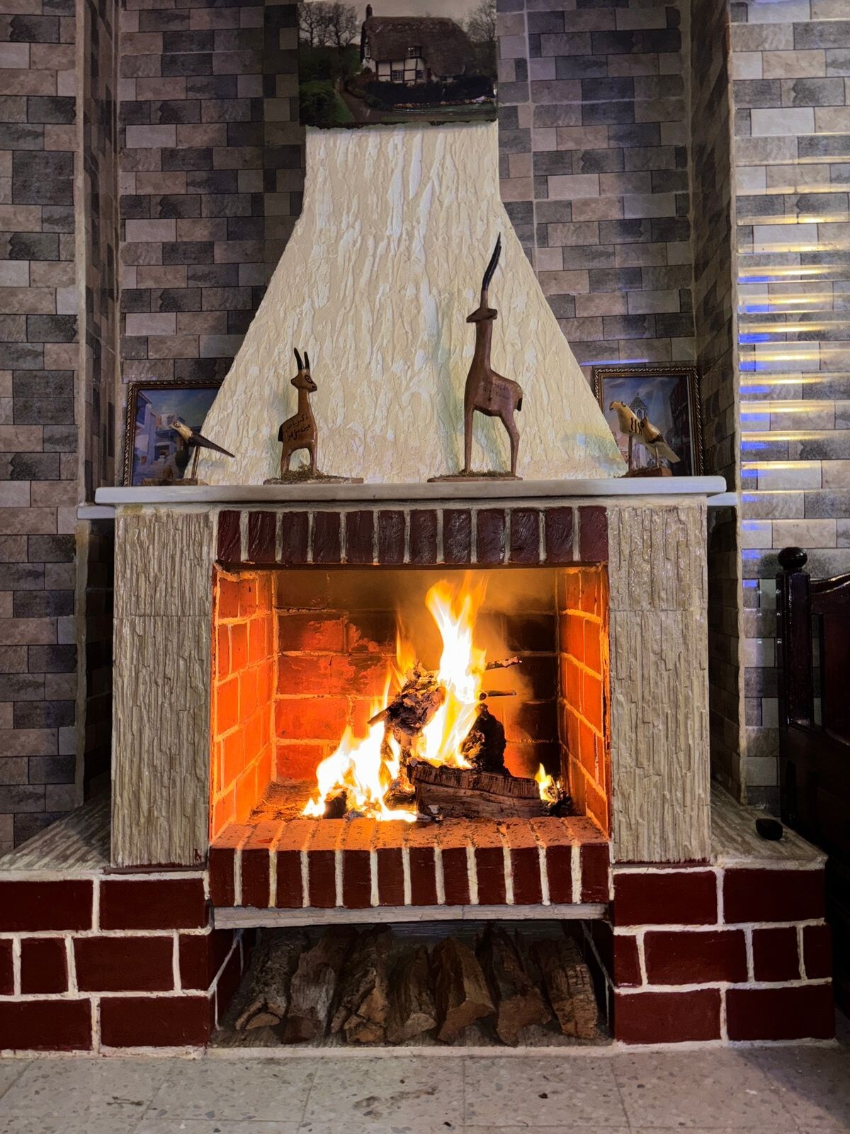 A traditional fireplace is prominently featured, with a warm fire glowing inside a brick hearth. Above the mantel, decorative wooden sculptures are displayed, complemented by framed images on the textured wall. The overall ambiance suggests a cozy gathering space.