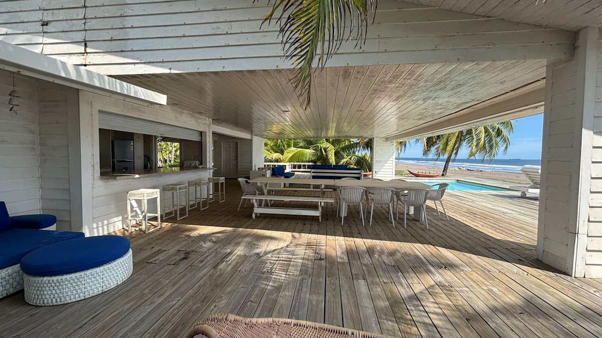 An open-air lounge area features comfortable seating arrangements, including a circular couch and multiple dining tables. A wet bar is positioned along one wall, while large windows provide views of the surrounding palm trees and the ocean beyond.