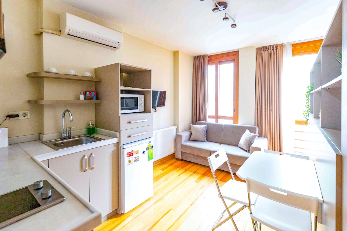 The open-concept studio features a kitchenette with modern appliances, including a refrigerator and microwave. A cozy sofa sits beside a window framed by light curtains, while a small dining table and chairs provide space for meals. Wooden flooring enhances the contemporary design.