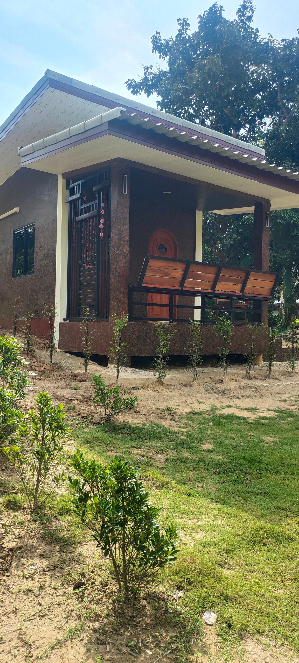 A single-story house with a brown exterior is shown, featuring a welcoming entrance that includes a wooden door. Surrounding the house, shrubs and small green plants are arranged in a neat garden area, with an open porch offering a view of the serene landscape.