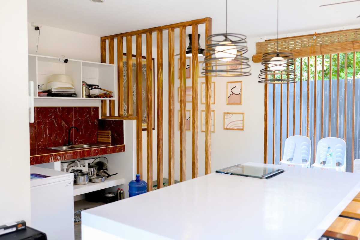 A modern kitchen area is showcased with a sleek white countertop and wooden accents. A stove is seen, alongside various cooking utensils organized on open shelving. Natural light fills the space, illuminating the decorative wall art and an adjacent seating area with light blue chairs.