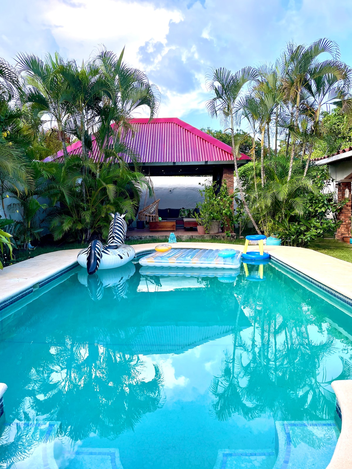 A tranquil swimming pool is framed by lush palm trees and tropical plants. A shaded bohio with a red roof is visible in the background. Colorful pool floats are arranged on the water's surface, reflecting the clear sky above.
