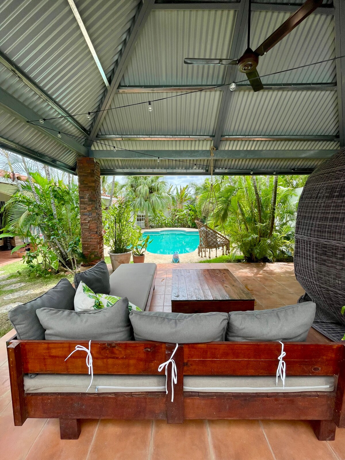 An outdoor seating area features a wooden sofa with gray cushions, positioned under a metal roof. Lush tropical plants surround the space, with a clear pool visible in the background. Natural light bathes the area, highlighting the inviting atmosphere.