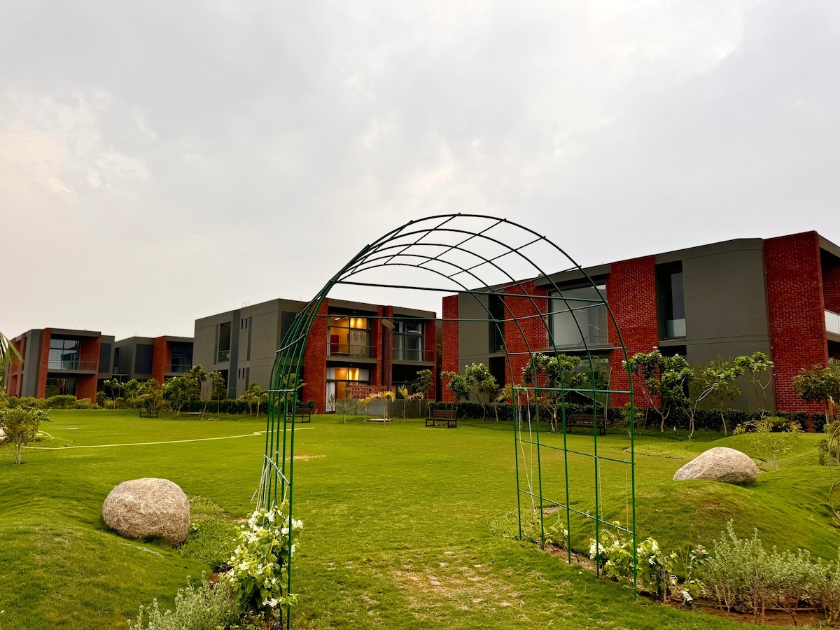 A green garden area is framed by a metal archway, leading to the modern buildings in the background. Lush grass extends throughout the space, complemented by large stones and neatly arranged shrubs.
