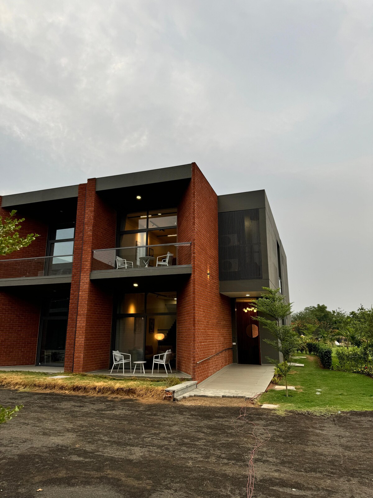 The exterior of a modern two-story villa is visible, featuring a combination of red brick and dark panels. A small balcony with two chairs overlooks a garden, while pathway lights softly illuminate the entrance. Lush greenery surrounds the building, enhancing its tranquil setting.