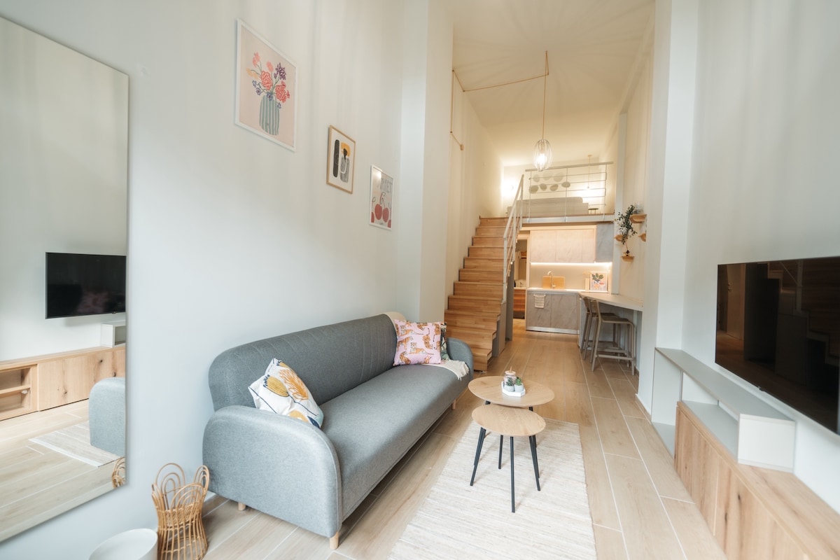 A contemporary living area is featured, showcasing a gray sofa adorned with decorative pillows. A small circular table sits in front, while a large mirror reflects the space. Natural light illuminates the area, highlighting the open staircase leading to the kitchen.