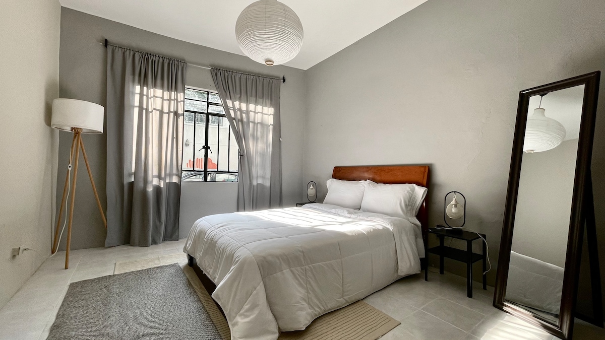 A spacious bedroom is observed with neutral-colored walls. A large window is covered by sheer curtains, allowing natural light to filter in. A wooden bed is positioned centrally, flanked by minimalist bedside tables with lamps. A full-length mirror stands against one wall, enhancing the room's openness.