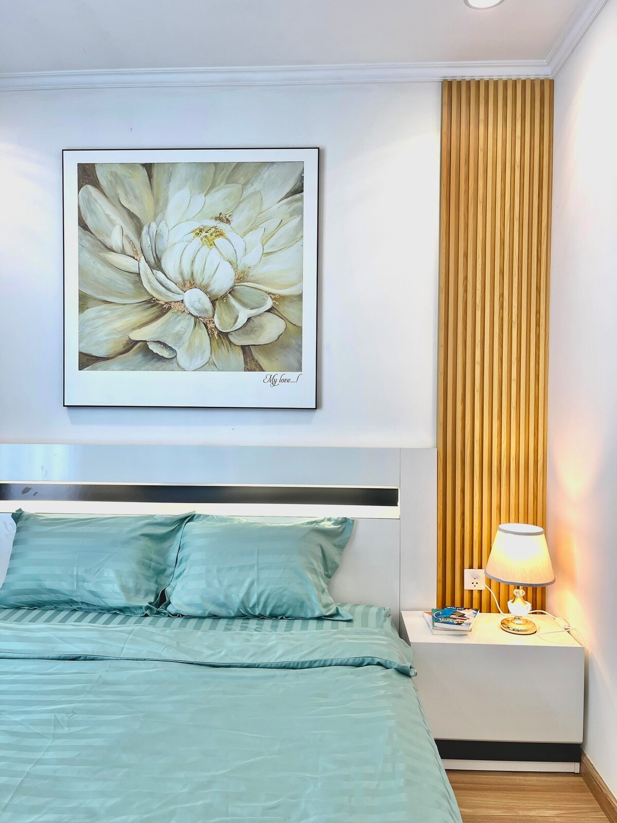 A spacious bedroom features a large bed with light teal bedding and matching pillows. A floral artwork adorns the wall above the bed. A bedside table includes a lamp and decorative items, while wooden wall panels add a touch of warmth to the bright environment.