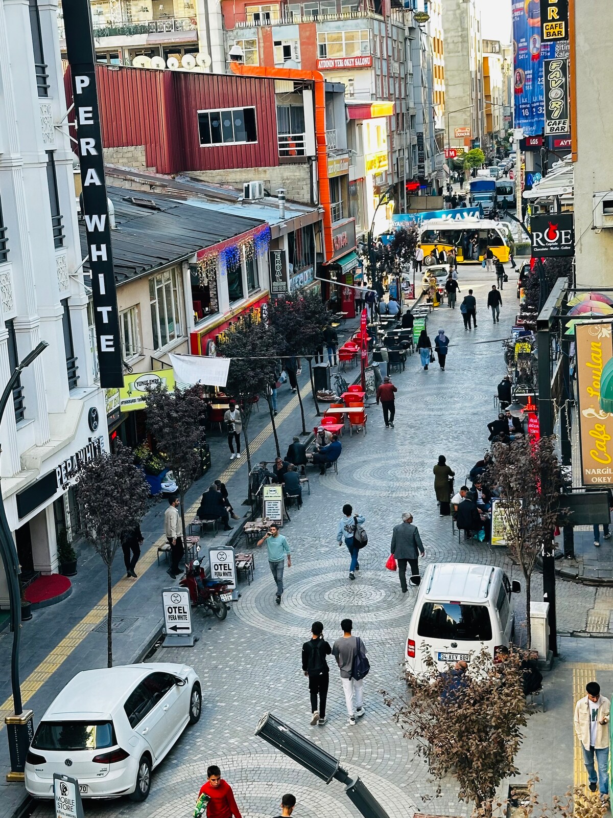 A bustling street scene showcases a vibrant atmosphere with pedestrians mingling among various shops and cafes. Cobblestone pathways are lined with colorful seating and storefronts. Vehicles are parked along the road, and trees add greenery to the urban landscape.