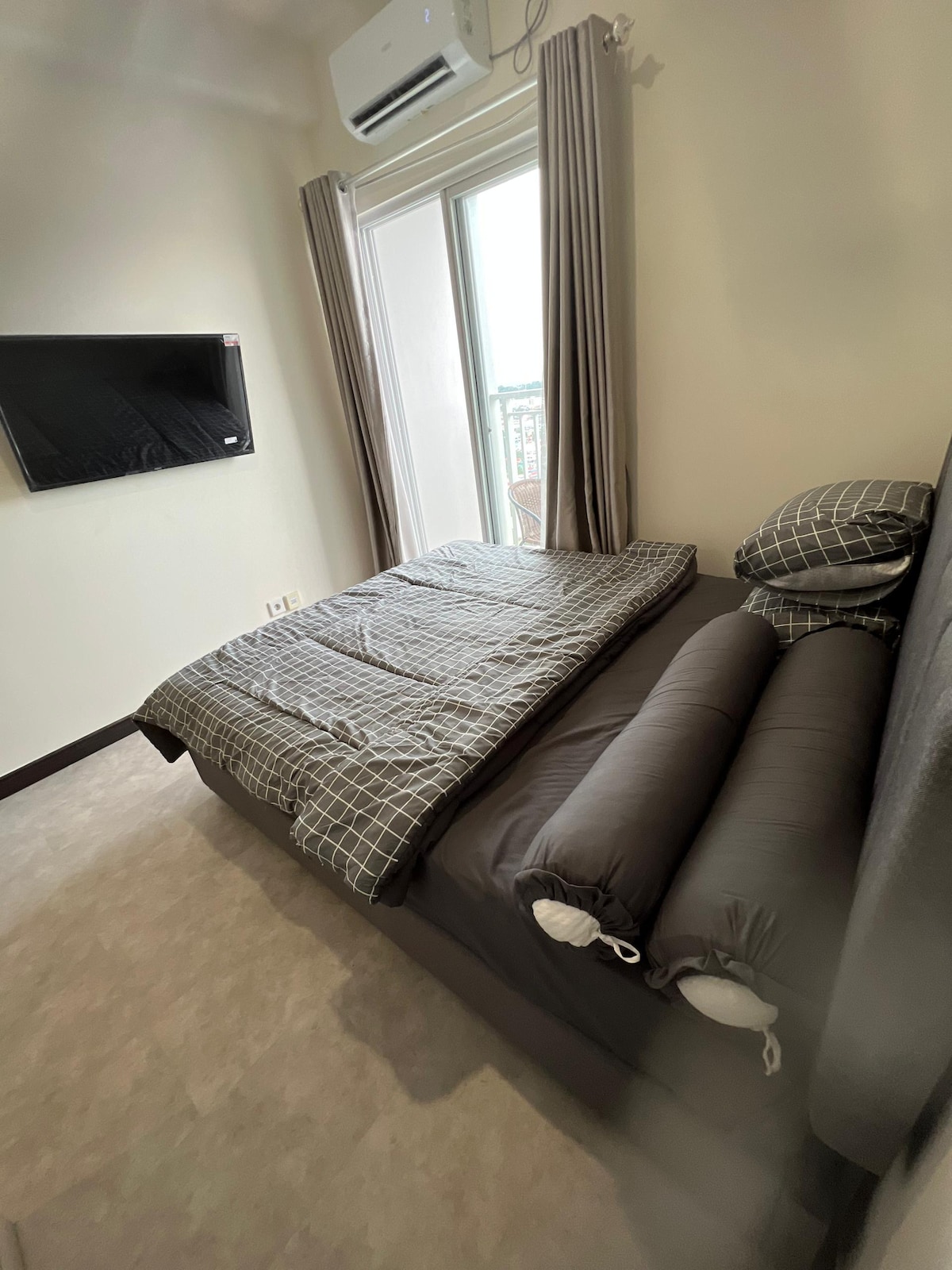 A modern studio bedroom is showcased, featuring a spacious bed dressed with a checkered duvet. Two decorative bolsters are positioned nearby. Natural light filters through floor-to-ceiling curtains. A wall-mounted television is visible across from the bed, creating an inviting atmosphere.