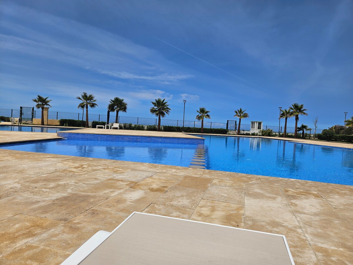 A large outdoor pool is bordered by smooth stone tiles, reflecting the clear blue sky. Palm trees line one edge of the pool area, adding a tropical touch to the serene environment. A shallow section can be seen, providing a gradual entry into the water.