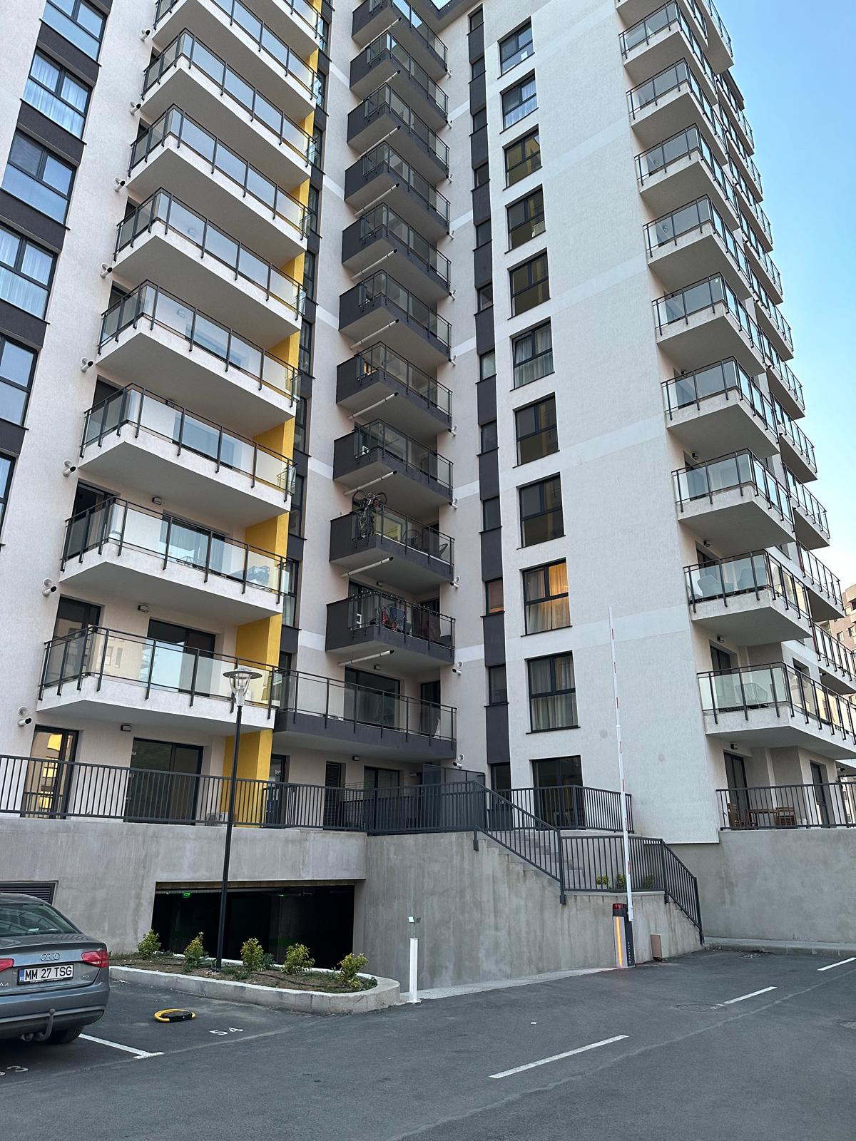A modern apartment building is depicted, featuring multiple floors with balconies that extend outward. The structure displays a clean façade with a mix of white and yellow accents. A parking area with a vehicle and landscaped planters is visible in the foreground.