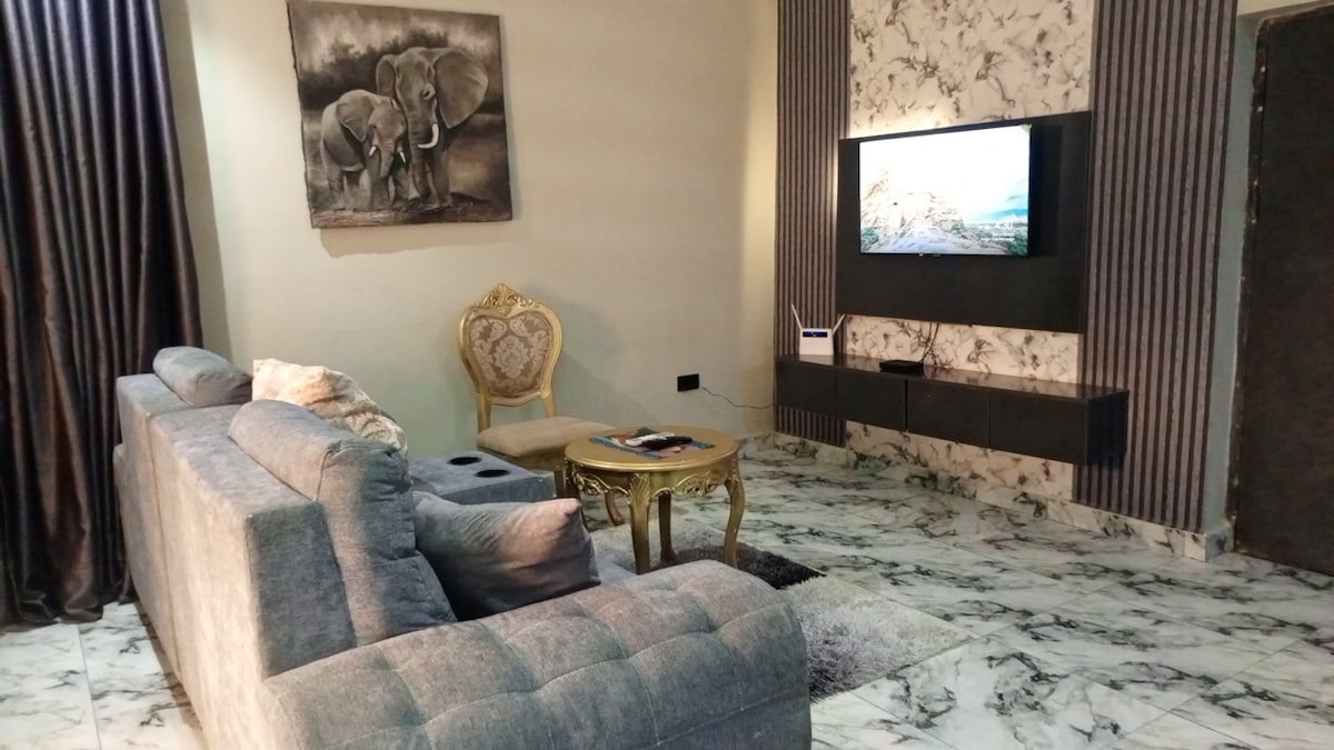 A living area features a plush gray sofa and a decorative gold-accented chair. A sleek coffee table rests in front of the seating. Wall-mounted artwork depicting elephants adds an earthy touch, while a television sits above a modern console, all on a marbled floor.