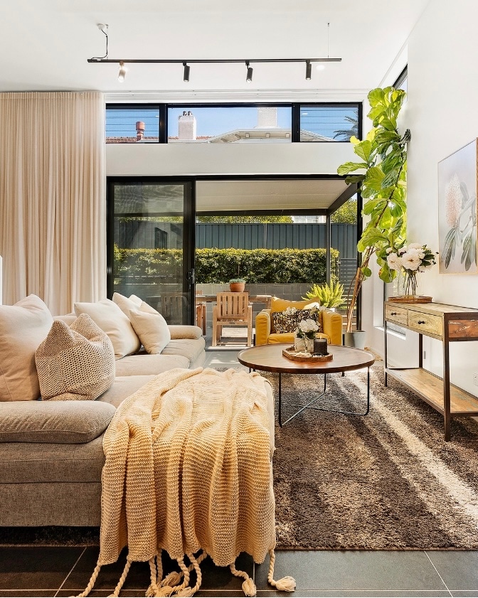 A comfortable lounge area features a plush sofa adorned with textured cushions and a knitted throw. A round coffee table sits centrally, complemented by decorative items. Large windows allow natural light to fill the space, with greenery visible outside, adding a refreshing touch.