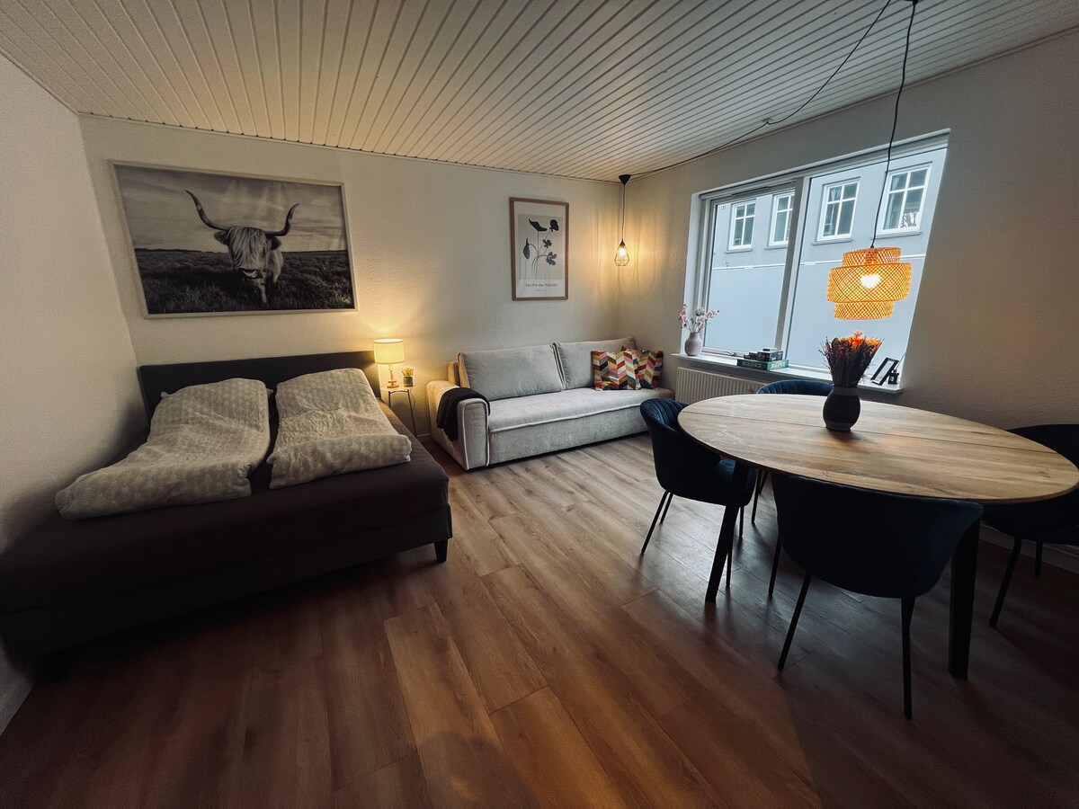 A welcoming living area is presented, featuring a spacious sofa bed and a decorative couch adorned with colorful cushions. A round dining table with dark chairs occupies one corner, while a large window allows natural light to brighten the space, complemented by soft lighting fixtures.