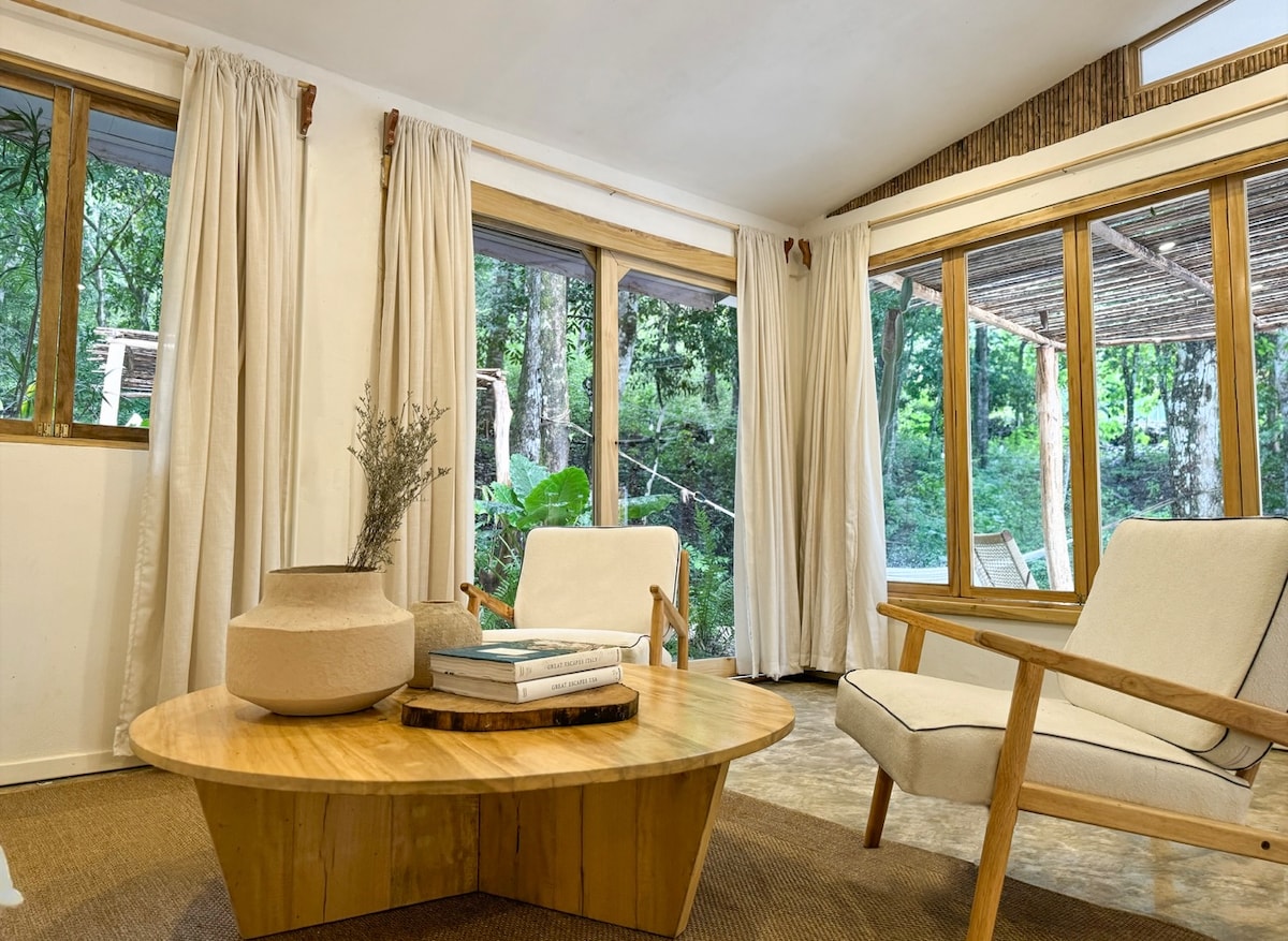A sunlit seating area features a circular wooden table surrounded by two minimalist armchairs. Natural light streams in through large windows, offering views of lush greenery outside. Soft, neutral curtains add an element of warmth, enhancing the room's connection to nature.