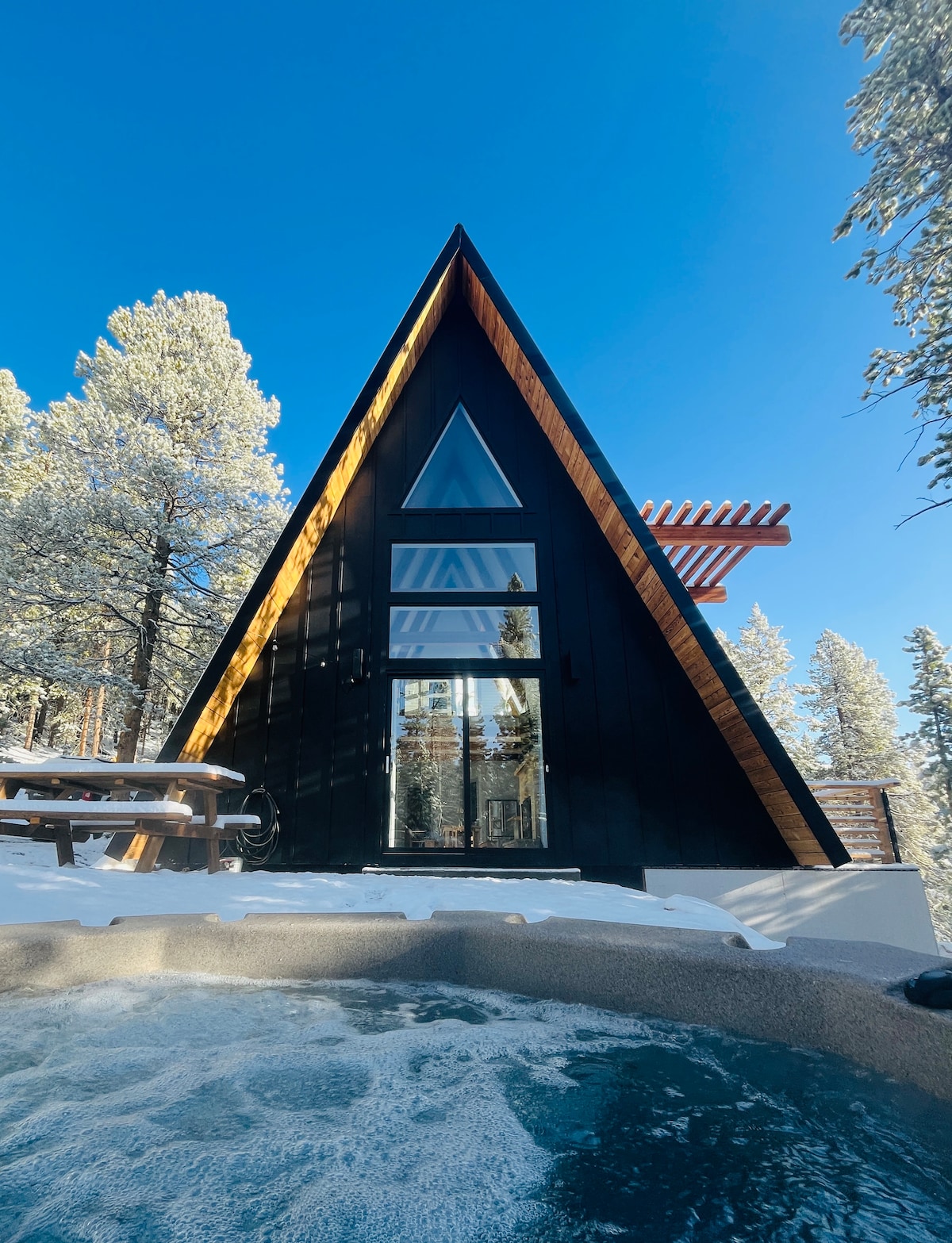 An A-frame cabin with a striking triangular design is surrounded by snow-dusted evergreens. A hot tub sits in the foreground, creating a relaxing atmosphere. Large glass doors and windows reflect the blue sky, emphasizing the cabin's blend with nature.