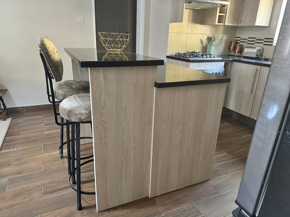 A modern kitchen is presented with a sleek island featuring a black countertop. Two barstools with plush seats are positioned at the island. The walls are adorned with light cabinetry, and a gas stove is visible in the background.