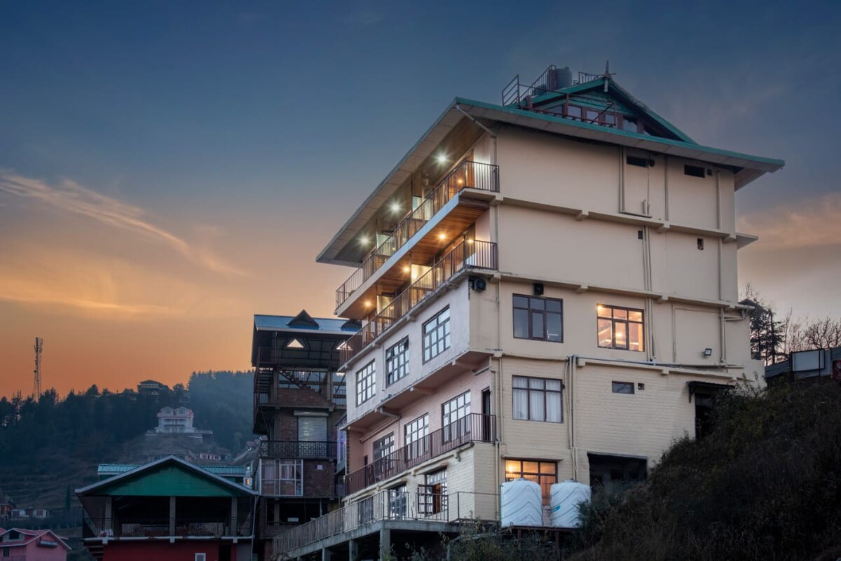 A multi-story building is captured at dusk, showcasing a mixture of modern and traditional architectural elements. Large windows reflect the soft colors of the sunset, while the rooftop features a distinctive green shelter. Surrounding hills are visible in the background.