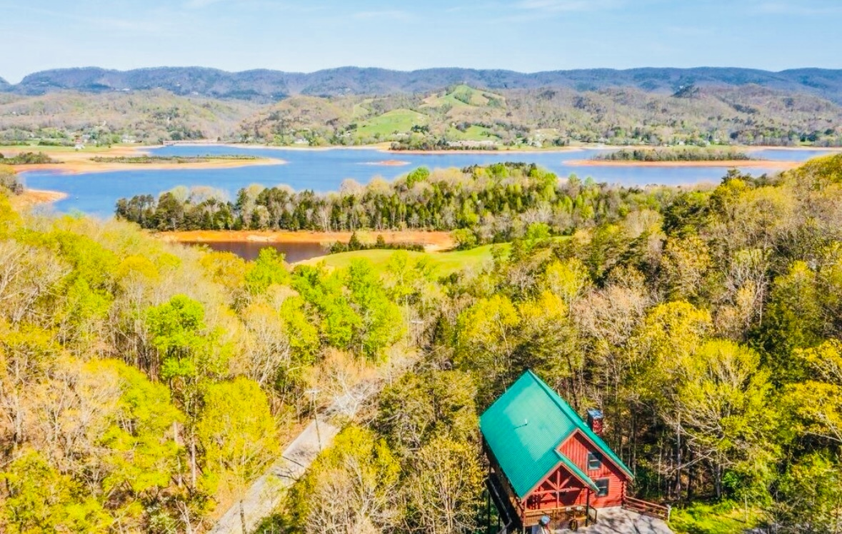 An aerial view captures the modern log cabin nestled among lush trees, with a green roof complementing the natural surroundings. Cherokee Lake glistens in the distance, framed by the rolling hills of Clinch Mountain, showcasing a serene and picturesque retreat.