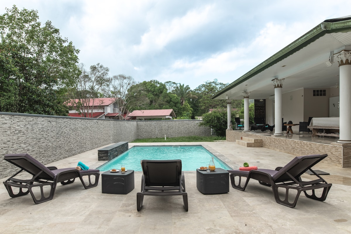 An outdoor space is depicted, featuring a private swimming pool surrounded by a stone terrace. Several lounge chairs are arranged around the pool, with a table holding refreshments nearby. Lush greenery can be seen in the background, adding a sense of privacy to the area.