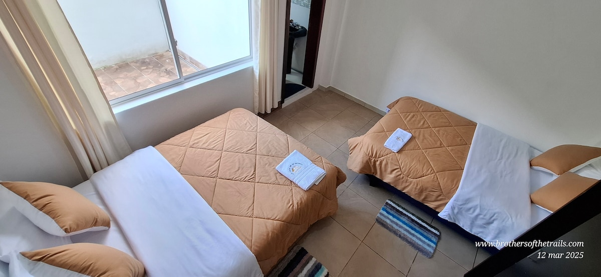 The room features two inviting beds, layered with soft beige bedspreads and neatly arranged white towels. Natural light filters in through a large window, illuminating the tiled floor and creating a calm environment. A small area rug adds a touch of color to the space.